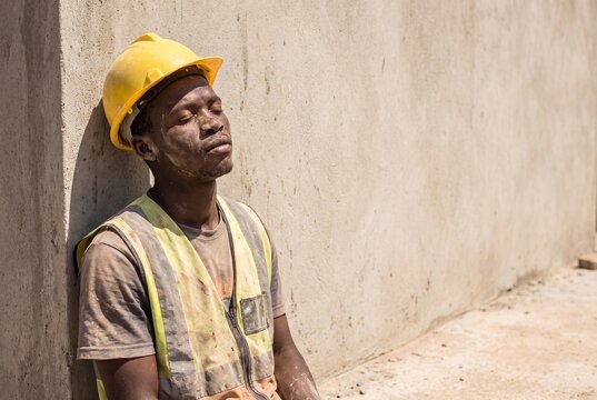 A tired African American construction worker covered in dust, wearing a yellow hard hat and safety vest, resting against a concrete wall on a sunny day.