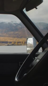 Vertical timelapse view from inside a car looking through the side window at a calm lake, autumn forest, and snowy mountain slopes under a cloudy sky. Vehicle interior frames the remote natural