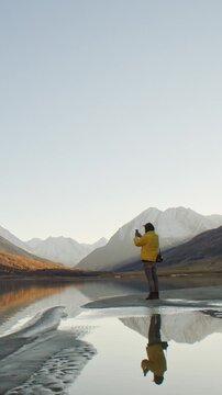 A man standing on the shore of a calm mountain lake takes a photo with his smartphone, surrounded by dramatic alpine scenery. Snow-capped peaks, autumn-colored slopes, and clear reflective water