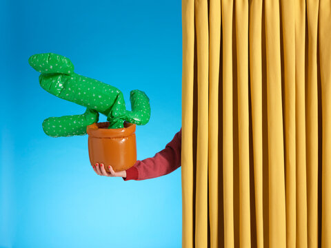 A hand holds a deflated blow up cactus in a pot from behind a curtain