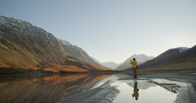 Traveler in a yellow jacket taking a photo with a smartphone while standing by a calm mountain lake at sunrise. Snow covered peaks, soft golden light, and mirror reflections create a scenic outdoor