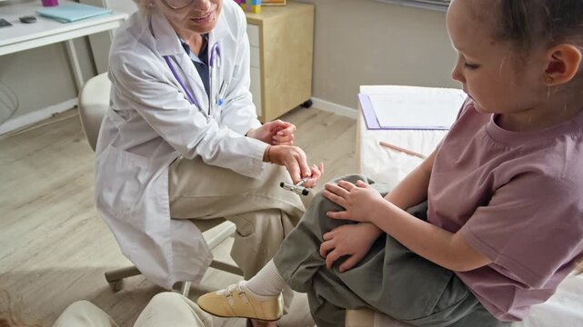 Caucasian female pediatrician checking Caucasian child knee reflex with neurological hammer in clinic. Footage ideal for pediatric healthcare marketing and medical services advertising