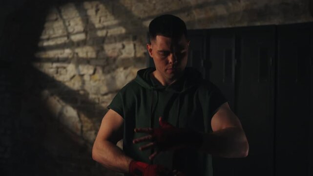 Focused young boxer wrapping hands with red bandages in dark gym. Male fighter preparing for workout or boxing match in locker room