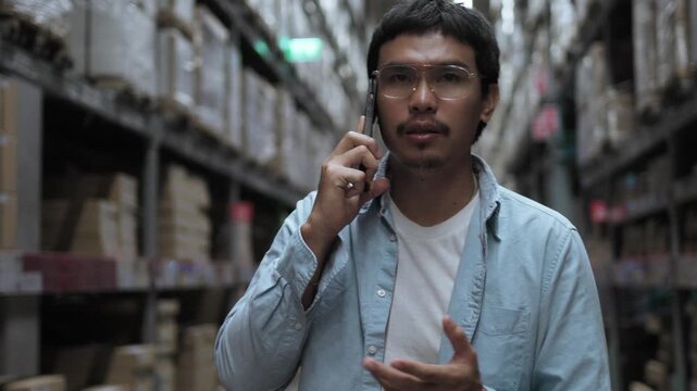 Asian Male Warehouse Worker Talking on Phone with Confident Gesture. Active Communication in Distribution Center