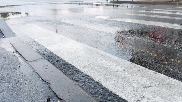 Deep puddles submerge parts of a white zebra crosswalk after a heavy downpour. The shimmering, wet asphalt captures a lonely, peaceful, and somber mood on the vacant urban road.