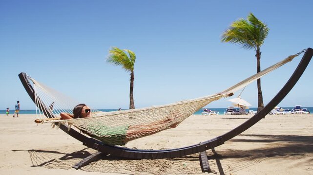 Summer vacation woman lying down on beach hammock relaxing sunbathing under the tropical sun resting on patio furniture swing bed at resort. Asian young adult. Isla Verde Beach in Puerto Rico.