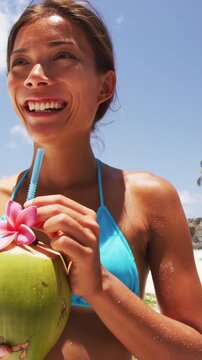 Vertical video. Cute Asian girl on beach vacation drinking fresh coconut water from a green coconut fruit cut from a palm tree. Healthy lifestyle woman eating local food on Caribbean travel holiday.