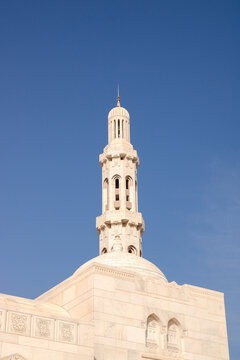 View of the ornate white stone minaret of the Sultan Qaboos Grand Mosque reaching into a clear blue sky with intricate architectural carvings in Muscat, Muscat Governorate, Oman.