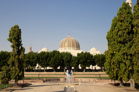 View of the Sultan Qaboos Grand Mosque with its golden dome and white architecture framed by lush green trees and a fountain under a clear blue sky in Muscat, Muscat Governorate, Oman.