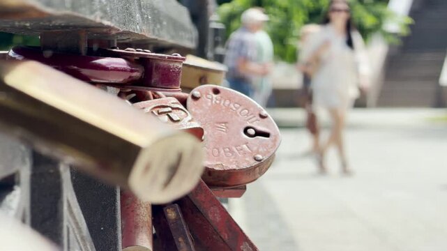 A rusty heart-shaped wedding lock clings to a metal railing. Blurry people enjoy a sunny day in the background. This sweet tradition evokes a deep feeling of romantic hope.