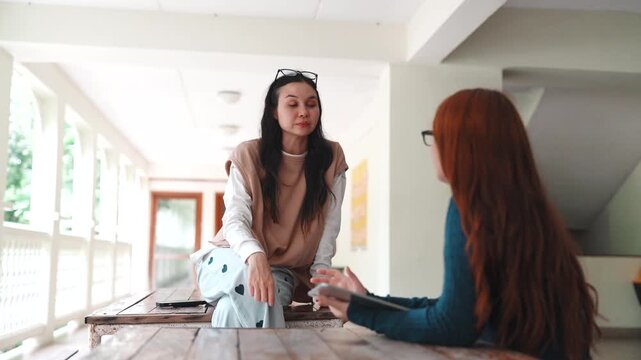 Two women students talking face to face at wooden table on campus. Casual friends having a serious discussion with tablet in university hallway.