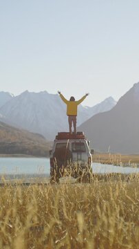 Traveler in a yellow jacket standing on the roof of an off road SUV with raised arms by a mountain lake in a remote alpine valley. Snowy peaks, golden grass, and bright sunlight create a powerful