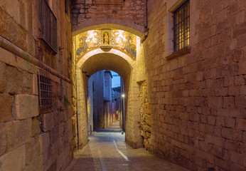 Obraz premium Narrow medieval street passing through an illuminated stone archway at dawn in the historic center of Girona, Catalonia, Spain.
