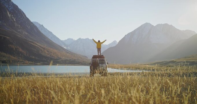 Traveler in a yellow jacket standing on the roof of an off road SUV with raised arms by a mountain lake in a remote alpine valley. Snowy peaks, golden grass, and bright sunlight create a powerful