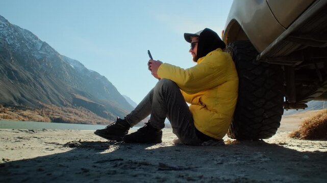 Male traveler in a yellow jacket using a smartphone while sitting on the ground and leaning against an off road SUV near a mountain lake. Scenic outdoor setting with alpine slopes, open shoreline, and