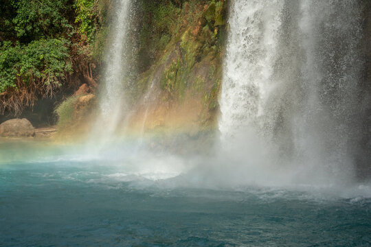 View of Havasu Falls with turquoise water and a rainbow appearing in the mist against a rocky cliff and green foliage in Supai, Arizona, United States.