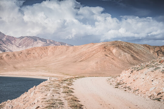 A dusty road on the Pamir Highway winds through mountain hills and slopes among rocks against the backdrop of the Tien Shan mountain peaks