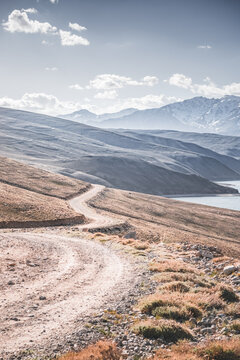A dusty road on the Pamir Highway winds through mountain hills and slopes among rocks against the backdrop of the Tien Shan mountain peaks