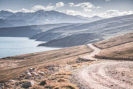 A dusty road on the Pamir Highway winds through mountain hills and slopes among rocks against the backdrop of the Tien Shan mountain peaks