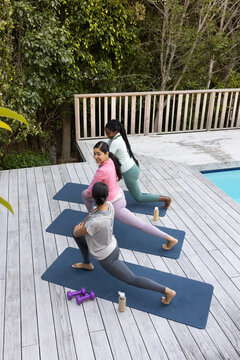 Diverse female friends practicing lunges on blue mats by pool with weights and bottles in leggings