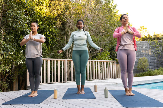 Diverse females practicing standing yoga in activewear on pool deck with dark mats, water bottles