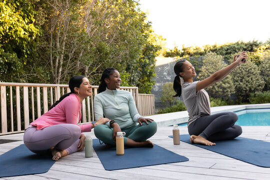 Diverse female friends in activewear sitting on yoga mats by pool deck holding smartphone, bottles