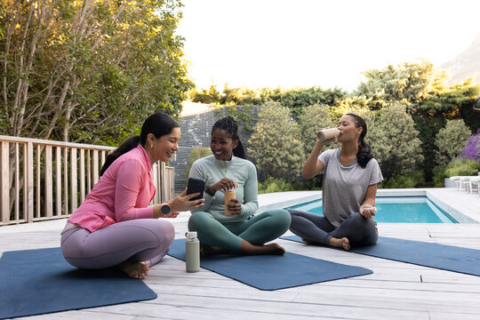 Diverse female friends sitting cross-legged on blue mats at pool in athletic wear holding bottles