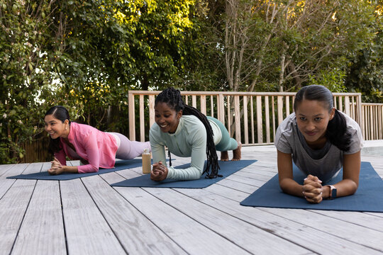 Diverse female friends performing forearm planks on wooden deck with dark mats and water bottle
