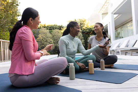 Diverse women in athletic wear sitting on blue mats at pool deck sharing smartphone, bottles near