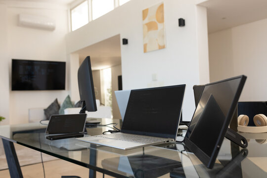 Silver laptop sitting open on glass desk in open-plan home office, beige headphones nearby