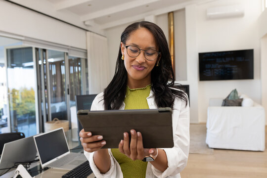 African American woman standing holding tablet wearing glasses in open-plan office with laptops