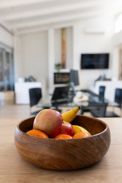 Wooden bowl of apples, bananas, mandarins is sitting on light wood table near sliding glass doors