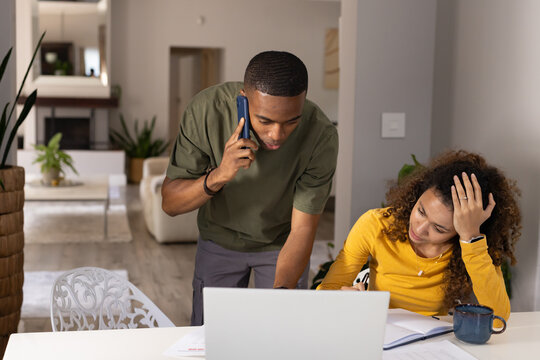 Diverse couple using open laptop at dining table, man leaning talking on phone, woman writing notes