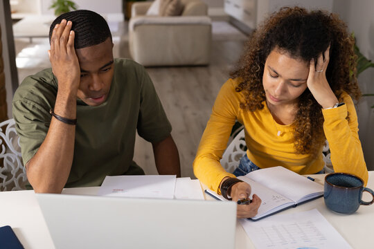 Couple sitting at dining table reviewing documents with laptop, notebook, pen and blue mug at home