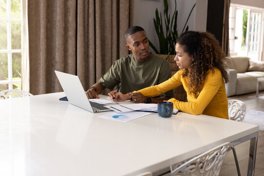 Couple sitting at home dining table, reviewing charts and using laptop, wearing olive, mustard tops