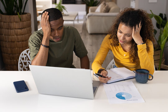Diverse couple reviewing bills on laptop at home, woman wearing mustard top writing notes