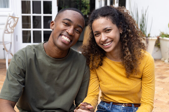 Couple holding hands, smiling on terracotta patio with French doors in olive tee and mustard top