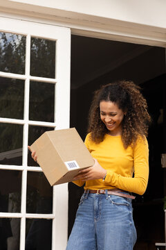 Adult female in yellow top, belt, holding box with barcode in doorway by white door, smiling