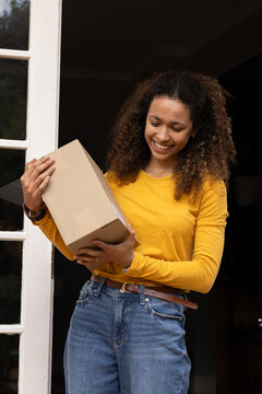 Adult woman standing in home doorway wearing mustard-yellow top and jeans, holding cardboard parcel