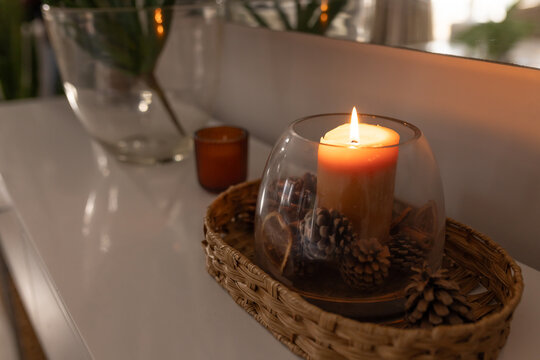 Pillar candle glowing under clear dome on wicker tray with pinecones and orange slices, near mirror