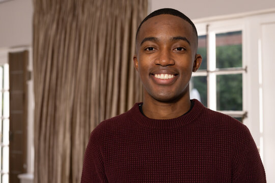 African man smiling and standing facing window inside home with maroon knit sweater and curtains