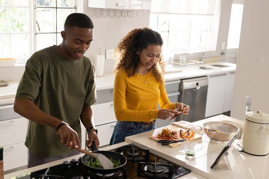 Diverse couple stirring green beans and peeling veggies at central island in modern kitchen, tablet