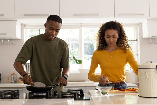 Diverse couple cooking at kitchen island, man using spatula on stove, woman chopping herbs
