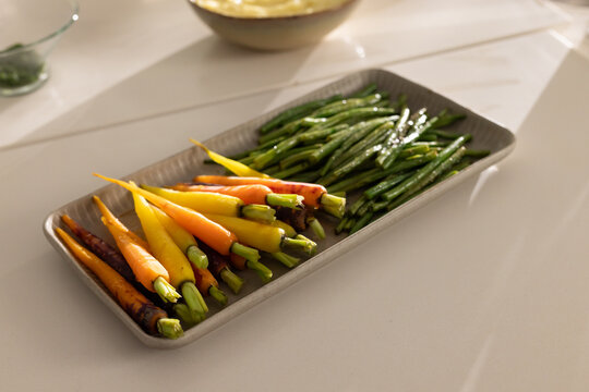 Platter is holding glazed stemmed baby carrots and green beans on light countertop, glass herb bowl