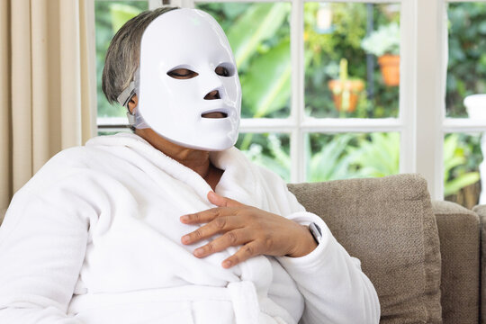 Senior African American woman sitting at home on sofa in white bathrobe, LED mask, watch visible
