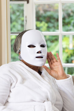 Senior African American woman sitting by paned window wearing LED mask and terry robe raising hand