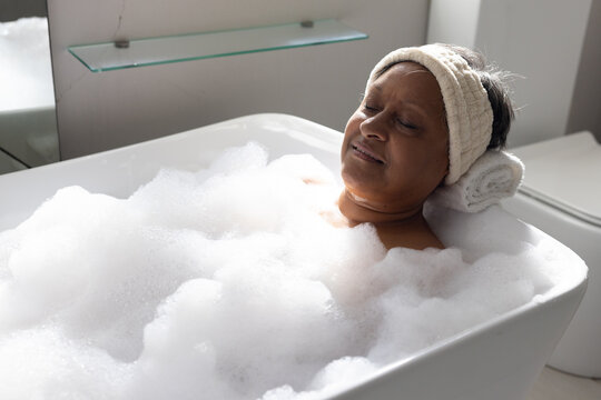 Senior African American woman reclining in bathroom tub, soaking in foam, rolled towel under head