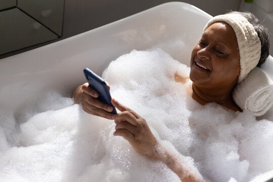 Senior African American woman reclining in tub with foam, using phone, headband, towel, copy space