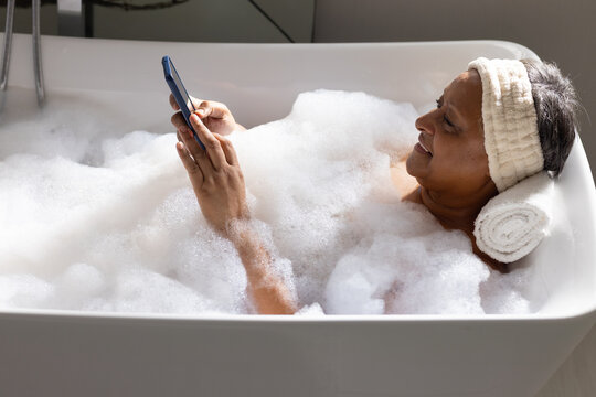 Senior African woman relaxing in tub in bathroom with foam, terry headband holding smartphone