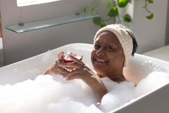 Mature adult African American woman reclining in foam bathtub at home holding red drink headband on